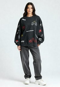 Felpa nera con grafiche e testo in bianco e rosso, inclusi fiamme e simboli. Abbinata a pantaloni grigi ampi e sneaker bianche.