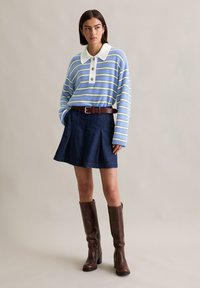 Pasiasty sweter w niebiesko-zielonych odcieniach z białym kołnierzykiem i trzema guzikami, zestawiony z ciemnoniebieską spódnicą dżinsową oraz brązowymi, wysokimi botkami na obcasie.