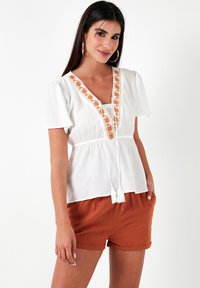 Witte blouse met korte pofmouwen, V-hals, oranje bloemenborduursel en een verstelbare taille. Gecombineerd met oranje shorts.
