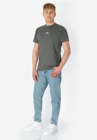 Man die in een ontspannen houding staat, gekleed in een donkergrijs t-shirt, lichtblauwe jeans en witte sneakers, tegen een effen witte achtergrond.