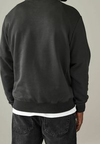 Sudadera negra con cuello redondo y puños acanalados, que presenta una textura suave. Llevada sobre una camisa blanca, se muestra desde atrás.
