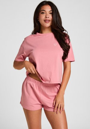 Frau mit langen dunklen Haaren trägt ein passendes rosa kurzärmeliges Shirt und Shorts und hebt das Shirt leicht an, um die Taille zu zeigen.