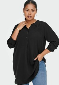 Maglietta nera a maniche lunghe con scollo henley e tre bottoni. Tessuto morbido, vestibilità comoda, orlo curvo. Indossata sopra dei jeans blu.