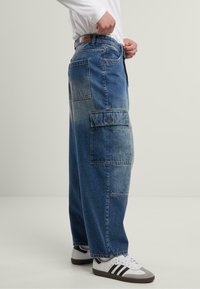 Baggy blue denim cargo pants with multiple pockets, faded texture, and a straight cut. Paired with white sneakers and a long-sleeve shirt.