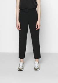 Svarta skräddarsydda byxor med bälte i midjan, raka ben och en slät yta, i kombination med grå och gula sportskor.