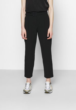 Pantalons noirs sur mesure avec une taille ceinturée, des jambes droites et une texture lisse, associés à des baskets de sport grises et jaunes.