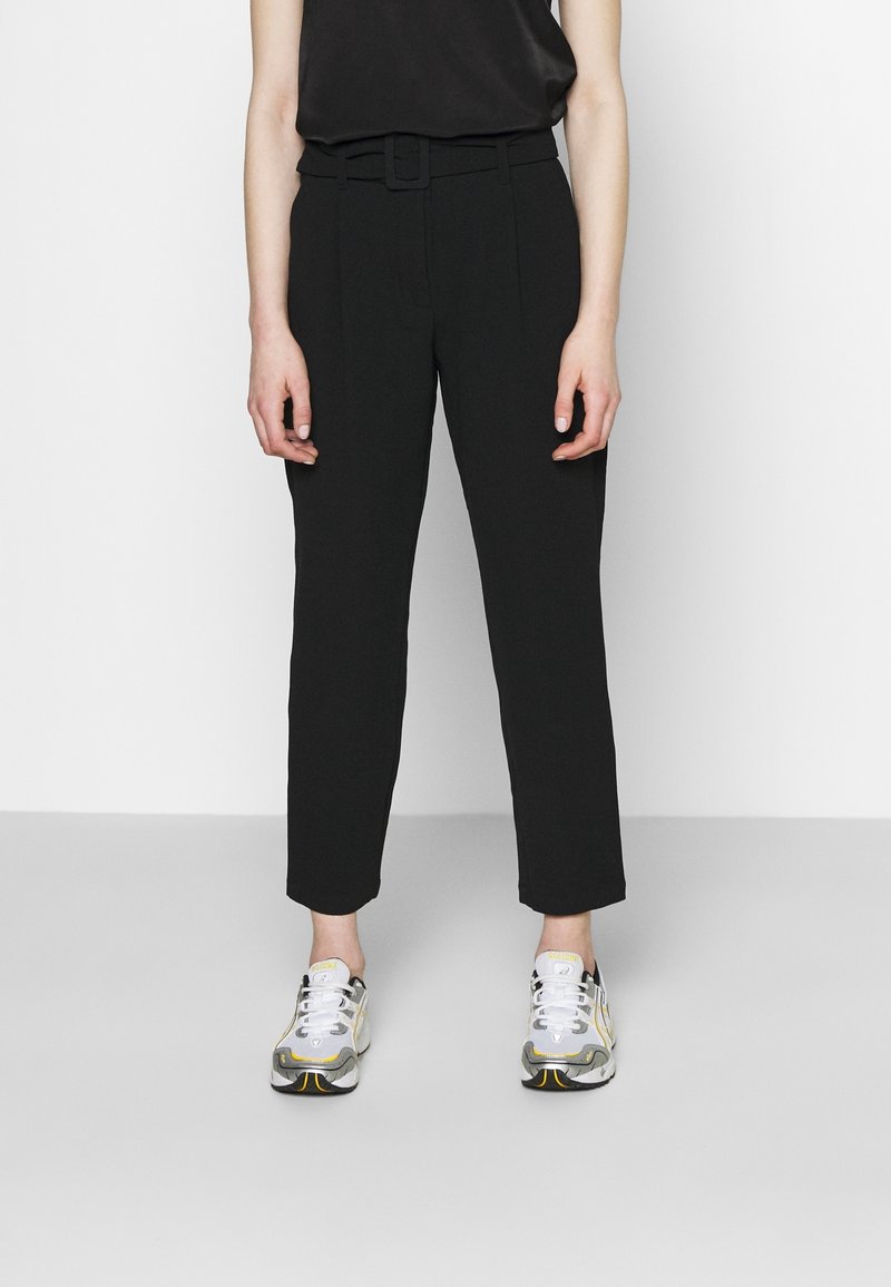 Svarta skräddarsydda byxor med bälte i midjan, raka ben och en slät yta, i kombination med grå och gula sportskor.