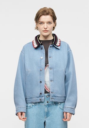 Donna con capelli corti che indossa una giacca di jeans azzurra con colletto lavorato a maglia colorato e jeans azzurri su uno sfondo semplice.