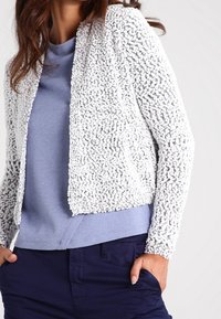 Cardigan blanc en maille texturée avec un devant ouvert et des manches longues, superposé à un haut bleu clair à manches longues, associé à un pantalon foncé.