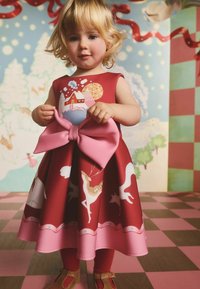 Czerwona sukienka dziecięca z dużą różową kokardą, zdobiona nadrukiem zimowej scenerii z zwierzętami i domami. Gładka w dotyku, o długości do kolan, z wzorzystym dołem.