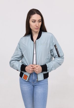 Woman with long brown hair wearing a light blue bomber jacket with black cuffs and light blue jeans, holding the jacket zipper.