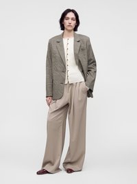 Femme portant un blazer à carreaux oversized, un top blanc boutonné, un pantalon beige large et des chaussures bordeaux, debout devant un fond uni.