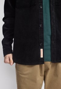 Zwarte corduroy overhemd met twee borstzakken, blauw-groene T-shirt eronder en kaki broek; met een label met de tekst "REVOLUTION."