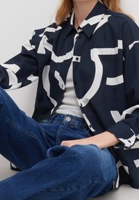 Chemise marine à boutons avec de grands motifs abstraits blancs, fabriquée en tissu léger. Associée à un jean en denim bleu taille haute.