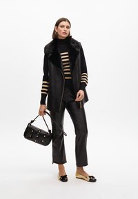 Chaleco de pelaje sintético negro con adornos de pelaje, sobre un suéter de rayas negras y doradas, combinado con pantalones de imitación de cuero negros, bolso negro y zapatos planos en dos tonos.