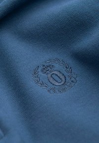 Kortsleeve, blå polotröja med texturerad tyg; har en broderad logotyp med ett krona och lagerkransdesign.