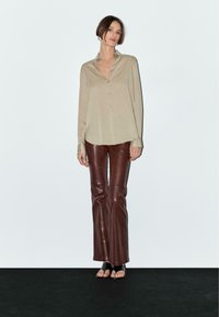Camicia beige con bottone e colletto, abbinata a pantaloni in pelle marroni a zampa d'elefante. Il modello si trova in piedi con le mani rilassate, mostrando uno stile aperto e informale.