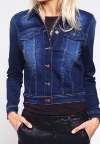 Donna che indossa una giacca di jeans scura aderente con bottoni, abbinata a una maglia nera e pantaloni in pelle nera, con le mani nelle tasche.