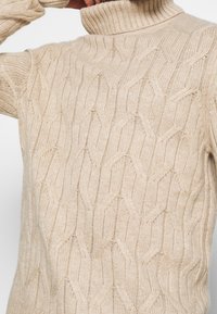 Beige stickad tröja med hög ribbad krage, prydd med ett intrikat bikaka-mönster och mjuk textur. Korta ärmar, figurnära silhuett.