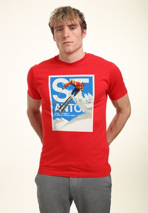 Joven con camiseta roja con gráfico de esquí y pantalones grises, de pie con las manos detrás de la espalda sobre fondo blanco.