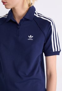 Polo bleu marine en coton, avec un col, une patte de boutonnage à trois boutons et des rayures horizontales blanches sur les manches. Logo Adidas affiché.
