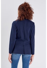 Blazer bleu marine à manches longues, design ajusté et détail d'ouverture à l'arrière. Confectionné dans un tissu lisse, porté avec un jean bleu.