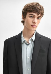 Zwarte op maat gemaakte blazer over een lichtblauw overhemd met knopen. De blazer heeft een gekartelde revers en een enkele borstzak.