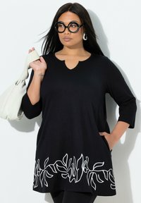 Schwarze Tunika mit 3/4-Ärmeln, ausgestattet mit einem gewellten Ausschnitt und weißer Blumenstickerei am Saum. Weicher Stoff mit lockerem Sitz.