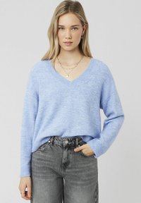 Pullover v-neck bleu clair en texture douce ; associé à un jean noir délavé taille haute. Les bijoux comprennent des colliers en or superposés et des boucles d'oreilles.