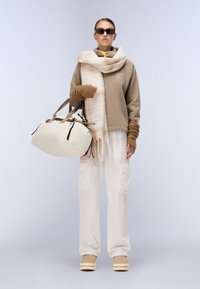 Felpa beige con cappuccio, sciarpa crema, guanti beige, pantaloni cargo bianchi e scarpe beige; porta una borsa da viaggio crema con dettagli neri.