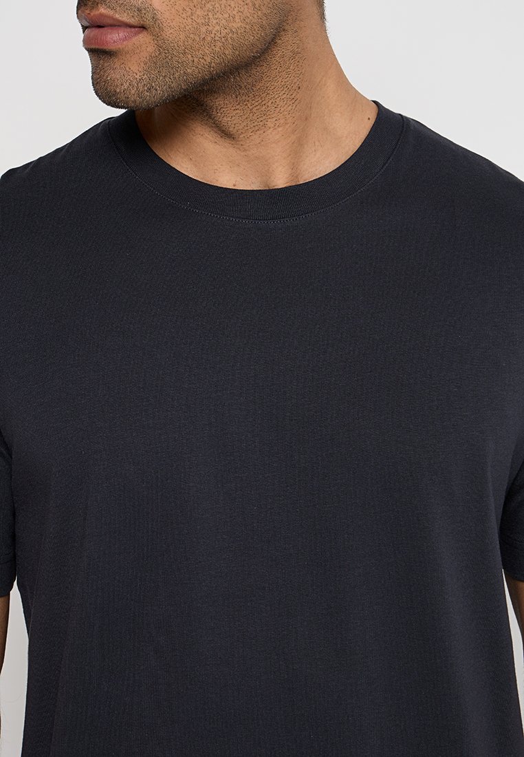 Zbliżenie mężczyzny w prostym czarnym t-shircie z okrągłym dekoltem, na którym widać część jego szyi, linii żuchwy i górnej części tułowia.