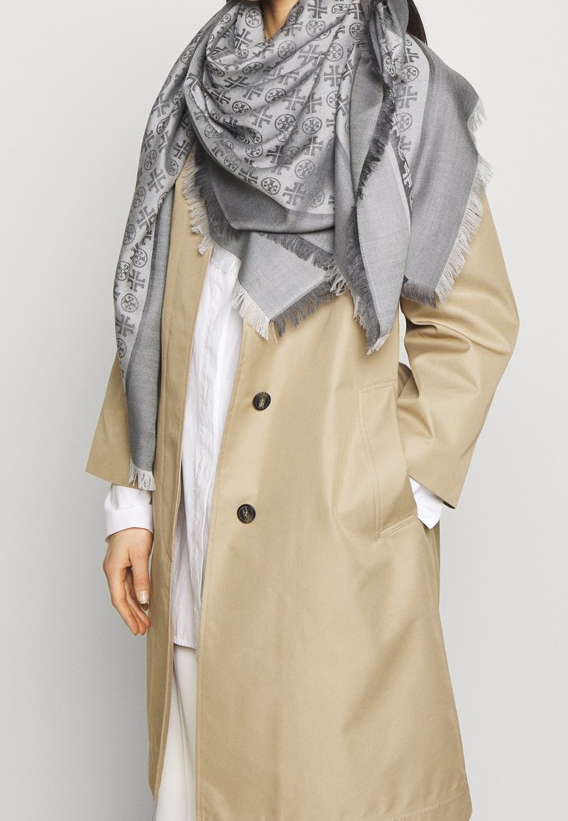 Trench-coat beige sur une chemise blanche et un pantalon, avec une grande écharpe grise à motifs enroulée autour du cou et des épaules.