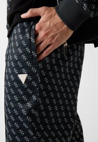 Schwarze gemusterte Hose mit Diamantdesign und weißen Logos. Enthält einen gebrandeten dreieckigen Aufnäher und Seitentaschen-Detail.