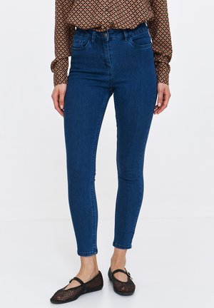 Cienkie niebieskie dżinsy o gładkiej fakturze, z wysokim stanem i minimalnymi detalami szwów, w połączeniu z czarnymi płaskimi butami z siateczki.