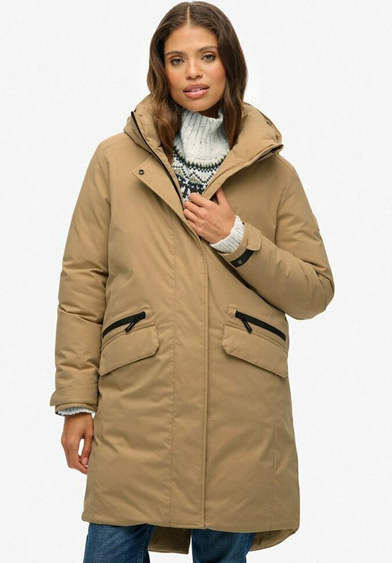 Beige gewatteerde jas met capuchon, voorzien van twee ritszakken aan de zijkant en een ritssluiting. Draagt over een gestructureerde trui, heeft een rechte pasvorm.