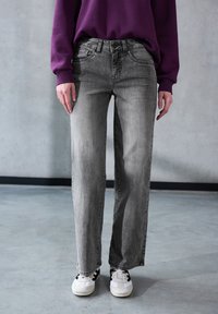Graue Jeans mit gerader Beinform, ausgestattet mit einem klassischen Fünf-Taschen-Design, subtilen Verwaschungen und einem Knopfverschluss. Kombiniert mit weißen Sneakern.