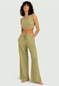 Donna con capelli ricci che indossa un top corto senza maniche di colore verde oliva e pantaloni larghi con coulisse, in piedi a piedi nudi con le mani nelle tasche.