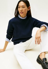 Navy textured knit sweater with a round neckline, layered over a light blue striped shirt, paired with white trousers and black loafers.