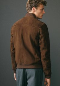 Chaqueta bomber de ante marrón con puños y dobladillo acanalados, que presenta una textura suave y detalles cosidos en la parte posterior.