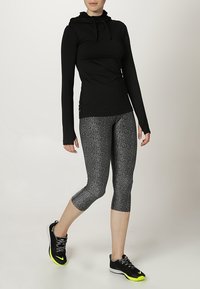 Sudadera negra de manga larga con capucha ajustable, combinada con leggins capri grises con un patrón moteado y zapatillas negras con acentos de neón.