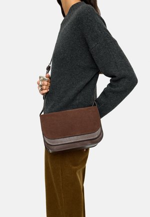 Osoba ubrana w ciemnoszary sweter i brązowe sztruksowe spodnie trzyma brązową skórzaną torbę na ramię z klapką zamykaną na zatrzask.