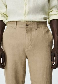 Pantalon en lin beige à coupe droite, doté d'une fermeture à boutons et de poches latérales. Le tissu présente une texture légère et texturée.