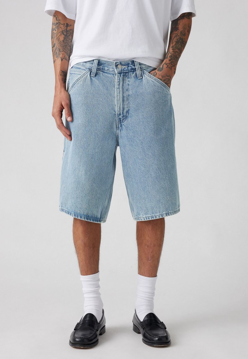 Pantalones cortos de mezclilla azul claro con un ajuste relajado, que presentan un cierre de cremallera, dos bolsillos frontales y un dobladillo deshilachado. Llevados con calcetines blancos y mocasines negros.