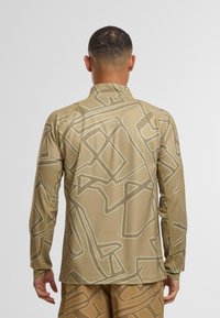 Langärmliges Oberteil in hellem Beige mit strukturiertem Stoff. Verfügt über ein abstraktes cremefarbenes Muster, einen hohen Kragen und dezente gelbe Akzente.