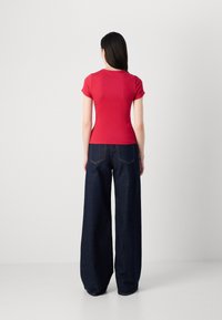 Une femme se tient dos à la caméra, portant un haut rouge ajusté et un jean bleu large, sur un fond gris neutre.