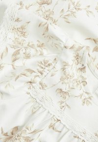 Weißer Stoff mit floralen Mustern und hellbraunen Akzenten, der eine Spitzenborte entlang des Ausschnitts und eine weiche, strukturierte Oberfläche aufweist.