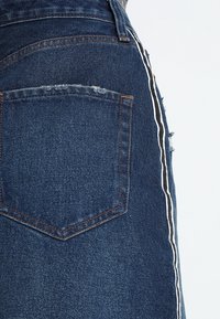 Falda vaquera de tela azul oscuro con un bolsillo trasero desgastado, franja lateral blanca y detalles de dobladillo deshilachado.