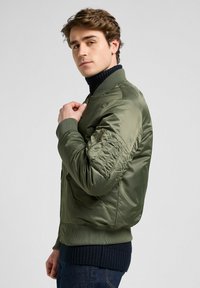 Giacca bomber verde oliva con chiusura a zip, polsini e orlo a costine, e una tasca con zip sul maniche sinistro. Finitura del tessuto liscia e lucida.