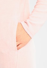 Tessuto rosa chiaro con una consistenza morbida, caratterizzato da una tasca laterale e un design a maniche lunghe con risvolto. È visibile un dettaglio di cucitura minimale.