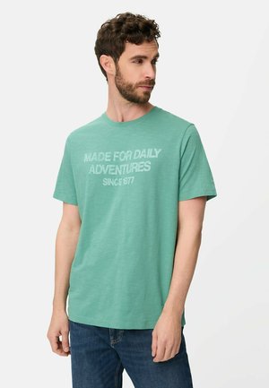 Mand med kort krøllet hår og skæg iført grøn T-shirt med teksten "MADE FOR DAILY ADVENTURES SINCE 1977" og blå jeans på hvid baggrund.